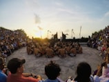 A large group of people is gathered outdoors in an amphitheater, watching a traditional performance at sunset. Performers, wearing patterned skirts, sit in a circle, engaged in a cultural dance. The spectators, dressed in casual and colorful attire, sit around the performers. Tall ceremonial poles and stone statues flank the stage, adding to the cultural atmosphere.