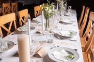 A long dining table is elegantly set with white tablecloths, featuring neatly arranged dinner plates, cutlery, and champagne glasses. The table is decorated with white candles and small floral centerpieces along with scattered golden confetti. Light wooden chairs are neatly aligned on both sides.