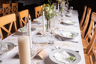 A long dining table is elegantly set with white tablecloths, featuring neatly arranged dinner plates, cutlery, and champagne glasses. The table is decorated with white candles and small floral centerpieces along with scattered golden confetti. Light wooden chairs are neatly aligned on both sides.