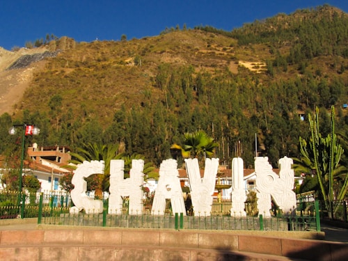 A sign with large white letters spelling 'Chavin' stands surrounded by manicured plants and small trees. In the background, a hillside covered with evergreens is visible under a clear blue sky. A lamp post with small ornamental lights is present on the left, and houses with red roofs are partially visible.