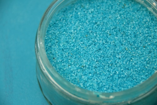 A clear glass container filled with numerous small blue granules, possibly sand or salt. The container is placed on a surface that matches the color of the granules.