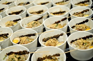 Multiple white bowls are arranged in rows, each containing a variety of food items. The contents include skewers with grilled meat, boiled eggs, and a mix of vegetables and herbs. Each bowl is served with a clear plastic fork.