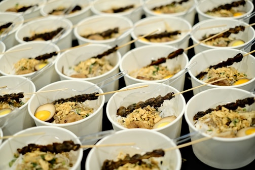 Multiple white bowls are arranged in rows, each containing a variety of food items. The contents include skewers with grilled meat, boiled eggs, and a mix of vegetables and herbs. Each bowl is served with a clear plastic fork.