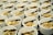 Multiple white bowls are arranged in rows, each containing a variety of food items. The contents include skewers with grilled meat, boiled eggs, and a mix of vegetables and herbs. Each bowl is served with a clear plastic fork.