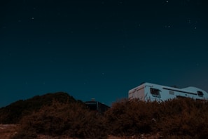 A camper van is parked on a hill under a clear, starry night sky. Bushes and shrubs are visible in the foreground, while the silhouette of the van is partly illuminated by moonlight.
