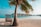 A serene tropical beach setting with a palm tree providing shade to a wooden chair on white sand. Overwater bungalows stretch into the turquoise sea under a clear blue sky.