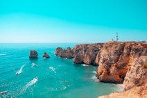A stunning coastal scene featuring rugged cliffs meeting a vibrant blue sea. Several boats create white trails in the water, adding movement to the peaceful setting. A lighthouse is perched on the highest cliff, enhancing the scenic beauty and providing a focal point.