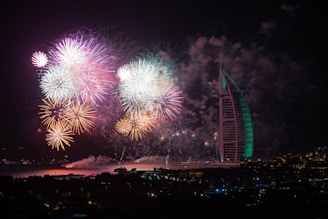 A spectacular display of colorful fireworks illuminates the night sky over a large, illuminated modern building with a sail-like structure by the waterfront. Multiple bursts of fireworks in hues of orange, pink, and green create a vibrant and festive atmosphere. The city skyline is dimly visible at the bottom, providing a silhouette against the bright explosions above.