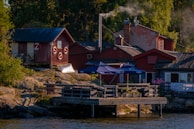 Wooden buildings with red paint and white trim are nestled among trees near a rocky waterfront. Smoke rises from a chimney, and life rings are attached to one of the buildings. Patio umbrellas are placed on a wooden deck by the water.