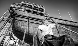 A black and white photograph depicts a man sitting in front of a large wooden boat under construction. The man appears to be taking a break, wearing a cap and a white shirt, with one hand on his forehead shielding his eyes and the other holding a cigarette. The boat is propped up by scaffolding and various wooden beams, suggesting an ongoing shipbuilding process.