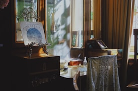 An elegant vintage interior with a polished wooden vanity table and mirror. A lace draped chair is in front of the table, which holds a decorative stand with a picture or placard featuring Cyrillic script. Sunlight streams through partially closed curtains, casting soft shadows and illuminating the room.