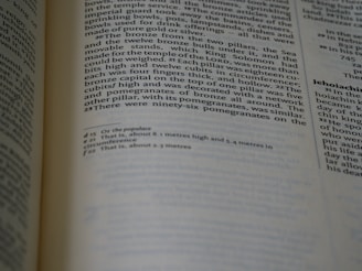 A close-up of an open book showing text in English, possibly a page from a religious or historical text, with two columns of text and footnotes at the bottom. The focus is on the left page.