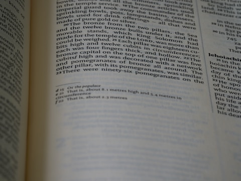 A close-up of an open book showing text in English, possibly a page from a religious or historical text, with two columns of text and footnotes at the bottom. The focus is on the left page.