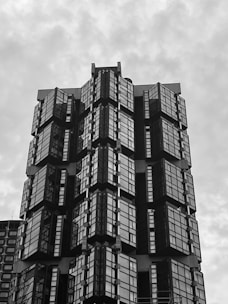 A high-rise building with a modern architectural design features a series of glass panes and sharp geometric shapes. The structure appears to have multiple protruding sections with reflective surfaces, creating a pattern of lines and angles.