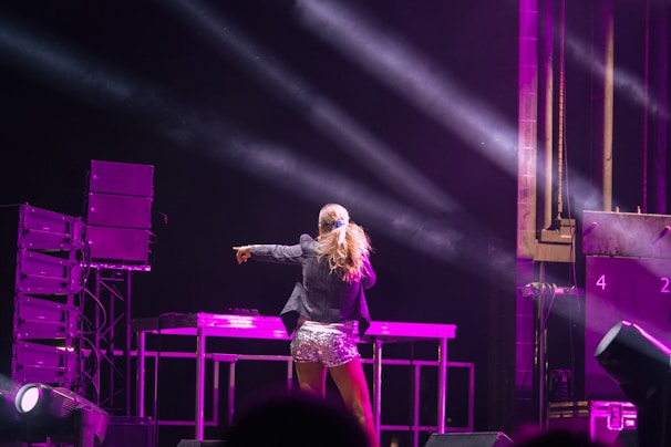 A performer is standing on stage under bright lights, facing away from the camera. They are wearing a dark jacket and sparkling silver shorts. Large speakers and stage equipment are visible around them, and beams of light illuminate the scene with a predominantly pink hue.