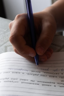 A close-up of a hand holding a blue pen, writing on lined paper. The handwriting is visible, with some text oriented horizontally.
