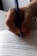 A close-up of a hand holding a blue pen, writing on lined paper. The handwriting is visible, with some text oriented horizontally.