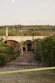 A rural setting with a quaint house partially obscured by lush greenery and plants. A concrete pathway leads to the entrance, flanked by well-maintained bushes and trees. In the background, rolling green hills extend into the distance under a light blue sky.