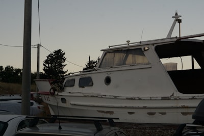 An old, weathered boat sits on land surrounded by several cars. The boat has peeling paint and rusty spots, indicating long-term exposure to the elements. A utility pole and some trees are visible in the background, with a clear sky above.