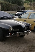A classic black car with a chrome bumper and round headlights is parked on a gravel surface. The license plate reads '500 KGX'. Next to it, there are a few more vehicles, including a beige hatchback. The background consists of a stone wall and some dense shrubs or bushes.