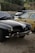 A classic black car with a chrome bumper and round headlights is parked on a gravel surface. The license plate reads '500 KGX'. Next to it, there are a few more vehicles, including a beige hatchback. The background consists of a stone wall and some dense shrubs or bushes.