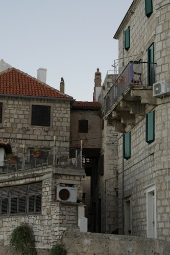 The image features a historic section of architecture with stone buildings and red tile roofs. The narrow alleyway between the buildings has a rustic charm. Wrought iron balconies and wooden shutters are prominent, and there's minimal greenery with a few potted plants on the ledge.