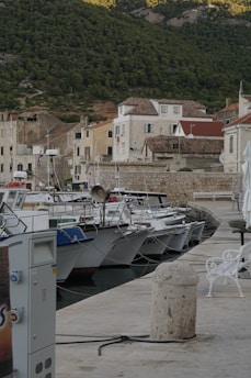 A marina with several small boats moored along a stone pier, surrounded by rustic buildings with terracotta roofs. Behind the buildings, lush green hills rise up, providing a natural backdrop. The scene is tranquil, hinting at a quaint seaside town.