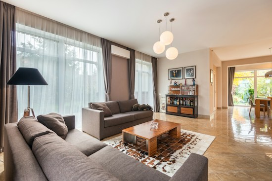 A modern living room with large windows allowing natural light to fill the space. The room is furnished with two gray sofas and a wooden coffee table on a patterned rug. A black floor lamp is situated next to one sofa. On the back wall, a wooden cabinet holds various items, and there are framed pictures on the wall above it. The room extends to a dining area with a glass table and chairs, visible through an open doorway. The decor is contemporary and elegant, creating an open and inviting atmosphere.