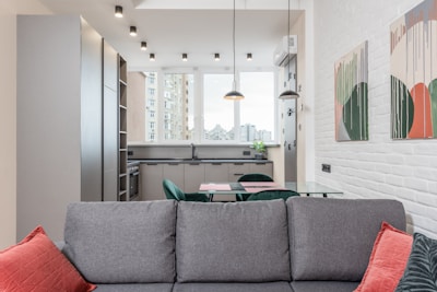 A modern and well-lit open-plan kitchen and living area featuring sleek grey cabinets, a glass dining table, and a comfortable grey couch with red cushions. Large windows offer a view of city buildings, while pendant lights hang from the ceiling. Contemporary abstract art adorns the white brick wall.