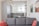 A modern and well-lit open-plan kitchen and living area featuring sleek grey cabinets, a glass dining table, and a comfortable grey couch with red cushions. Large windows offer a view of city buildings, while pendant lights hang from the ceiling. Contemporary abstract art adorns the white brick wall.