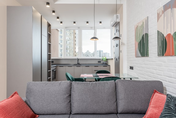 A modern and well-lit open-plan kitchen and living area featuring sleek grey cabinets, a glass dining table, and a comfortable grey couch with red cushions. Large windows offer a view of city buildings, while pendant lights hang from the ceiling. Contemporary abstract art adorns the white brick wall.