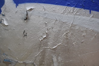 Peeling paint on a textured wall reveals an underlying layer beneath. The upper portion features a rough patch of blue paint, while the beige surface below is cracked and flaking.