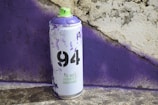 A spray paint can with a purple and light green design is positioned in front of a textured wall, which appears to have been sprayed with purple paint. The can is labeled with '94' and the phrase 'The Most Versatile Spraypaint'. The surface beneath the can is rough and mottled.