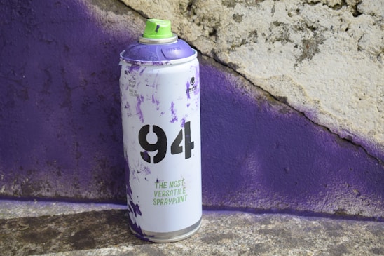 A spray paint can with a purple and light green design is positioned in front of a textured wall, which appears to have been sprayed with purple paint. The can is labeled with '94' and the phrase 'The Most Versatile Spraypaint'. The surface beneath the can is rough and mottled.