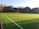 A well-maintained soccer field with vibrant green artificial turf, outlined by white lines. A single goalpost stands at one end of the field. The background features a dense row of trees under a clear blue sky, suggesting a serene natural setting.