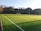 A well-maintained soccer field with vibrant green artificial turf, outlined by white lines. A single goalpost stands at one end of the field. The background features a dense row of trees under a clear blue sky, suggesting a serene natural setting.