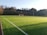 A well-maintained soccer field with vibrant green artificial turf, outlined by white lines. A single goalpost stands at one end of the field. The background features a dense row of trees under a clear blue sky, suggesting a serene natural setting.