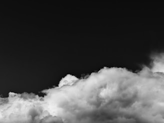 A dark sky contrasted by large, fluffy white clouds creating a dramatic effect. The image highlights the stark difference between the clear, shadowy expanse above and the textured, illuminated clouds below.