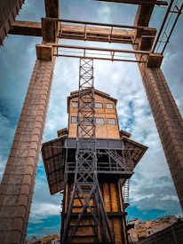 An industrial building with a tower structure composed of steel beams and brick walls, viewed from a low angle. The architectural design features geometric shapes and a framework of scaffolding. The sky is overcast with a mix of clouds, adding a dramatic effect to the scene.