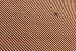 A person is wearing a yellow safety vest while working on a large, sloped roof made of brown and orange tiles in diagonal stripes.