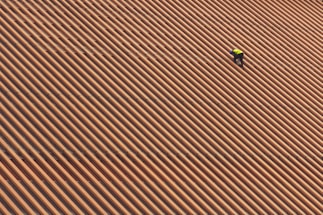 A person is wearing a yellow safety vest while working on a large, sloped roof made of brown and orange tiles in diagonal stripes.