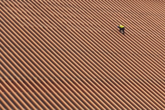 A person is wearing a yellow safety vest while working on a large, sloped roof made of brown and orange tiles in diagonal stripes.