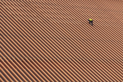 A person is wearing a yellow safety vest while working on a large, sloped roof made of brown and orange tiles in diagonal stripes.