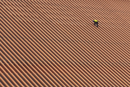 A person is wearing a yellow safety vest while working on a large, sloped roof made of brown and orange tiles in diagonal stripes.