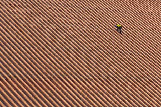A person is wearing a yellow safety vest while working on a large, sloped roof made of brown and orange tiles in diagonal stripes.