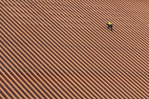A person is wearing a yellow safety vest while working on a large, sloped roof made of brown and orange tiles in diagonal stripes.