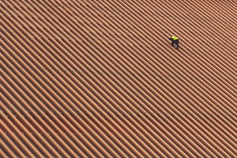 A person is wearing a yellow safety vest while working on a large, sloped roof made of brown and orange tiles in diagonal stripes.