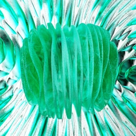 A central green, translucent structure resembling layered leaves or petals dominates the composition. Surrounding it is a textured background of swirling patterns in a mix of teal and white, evoking a sense of movement and depth.