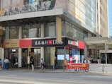 A street scene outside a commercial building named Emperor Commercial Centre. The building has signage for DBS Bank with a mix of English and Chinese characters. Several people are walking on the sidewalk, while others are standing near an entrance to the Hong Kong Station on Des Voeux Road Central. There is a large advertisement poster featuring multiple people near the entrance.