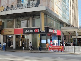 A street scene outside a commercial building named Emperor Commercial Centre. The building has signage for DBS Bank with a mix of English and Chinese characters. Several people are walking on the sidewalk, while others are standing near an entrance to the Hong Kong Station on Des Voeux Road Central. There is a large advertisement poster featuring multiple people near the entrance.